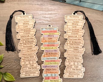 May include: Three light-colored wooden bookmarks shaped like stacks of books, each engraved with titles including The Shining, Misery, and Carrie. Two bookmarks feature black tassels at the top, while the center one has a small hole for hanging.