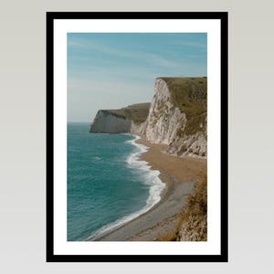 May include: A framed photograph of a coastal landscape. The image shows a turquoise sea, a sandy beach, and white cliffs with green vegetation under a blue sky. The photograph is presented in a black frame.