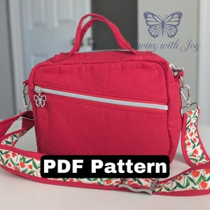 Puede incluir: Un bolso bandolera rojo con cremallera plateada y un charm decorativo de mariposa. El bolso tiene una correa estampada con flores rojas y hojas verdes. El texto "PDF Pattern" se muestra en la parte inferior.