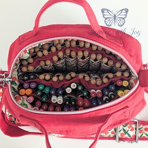 May include: A red canvas bag with a zipper, handle, and shoulder strap. The interior is divided into multiple compartments filled with various colored pens and wooden-topped tools. The lining features a floral pattern.
