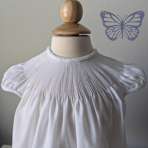 May include: A white, short-sleeved dress with a smocked bodice and a high neckline. The dress is displayed on a mannequin. A light purple butterfly graphic is on the wall behind the dress.