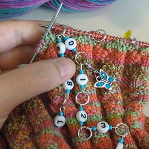 以下が含まれることがあります： 編み物のプロジェクトのクローズアップ。オレンジ、緑、ピンクの糸を使用。ステッチマーカーは、黒い数字の白いビーズで、シルバーのリングとターコイズビーズで接続されています。青い蝶のチャームも付いています。