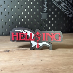 Könnte beinhalten: Ein 3D-gedrucktes Schild mit dem Wort HELLSING in fetten roten Buchstaben. Der Buchstabe O ist durch ein stilisiertes rotes Augensymbol mit einer tropfenförmigen Form darunter ersetzt. Ein weißes, gezacktes, flügelartiges Element liegt dahinter.