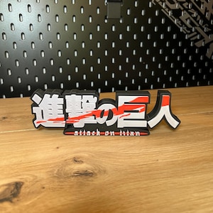Könnte beinhalten: Ein dreidimensionales Attack on Titan-Logo in Schwarz, Weiß und Rot, mit japanischen Schriftzeichen und dem englischen Text "attack on titan". Das Logo steht auf einer Holzoberfläche.