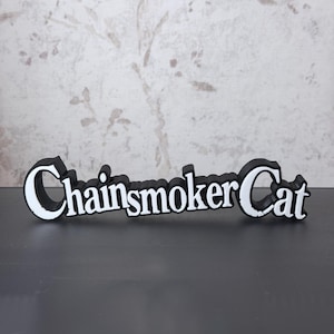 Könnte beinhalten: Ein dreidimensionales Schild mit dem Schriftzug "ChainsmokerCat" in einer fetten, stilisierten Schriftart. Die Buchstaben sind weiß mit einer dicken schwarzen Umrandung und einem schwarzen Seitenprofil, was einen Schichteffekt erzeugt.