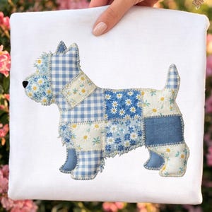 May include: A white square fabric with a patchwork dog design. The dog is composed of various blue and white patterned fabrics, including gingham, floral, and solid denim. The dog has a black nose and is held by a person.