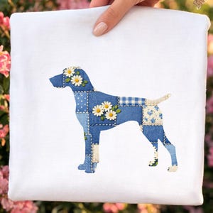 May include: White square fabric featuring a patchwork dog design. The dog silhouette is composed of various blue denim patches, adorned with white daisy accents. The design is held by a hand, with a blurred floral background.