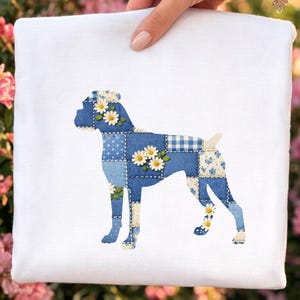 May include: White square fabric with a Boxer dog silhouette. The dog is made of blue denim patchwork with white daisy accents. The fabric is held by a hand, with pink flowers in the background.
