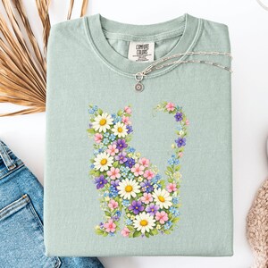 May include: A light green t-shirt featuring a floral cat design. The cat is composed of white daisies, pink, purple, and blue flowers. The shirt has a crew neck and a tag that says "Comfort Colors". A silver necklace with a sun pendant is on the shirt.