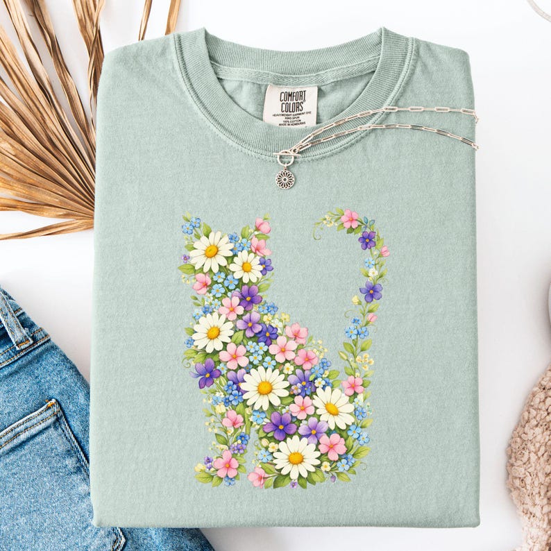 May include: A light green t-shirt featuring a floral cat design. The cat is composed of white daisies, pink, purple, and blue flowers. The shirt has a crew neck and a tag that says "Comfort Colors". A silver necklace with a sun pendant is on the shirt.
