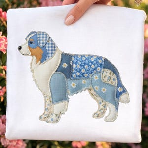 May include: A white square textile featuring an appliqued dog design. The dog is a patchwork of blue fabrics, including gingham, floral, and solid patterns. The dog's fur is white and beige, with a detailed embroidered outline.