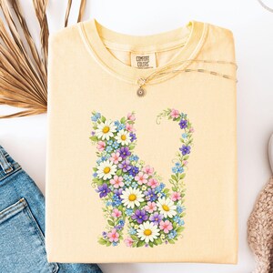 May include: A light yellow sweatshirt featuring a floral cat design. The cat silhouette is composed of white daisies, purple, pink, and blue flowers. A delicate gold necklace with a sun pendant rests on the sweatshirt.