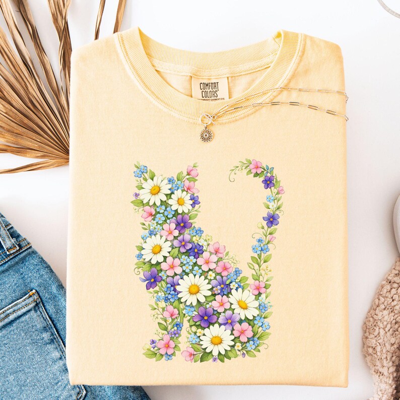 May include: A light yellow sweatshirt featuring a floral cat design. The cat silhouette is composed of white daisies, purple, pink, and blue flowers. A delicate gold necklace with a sun pendant rests on the sweatshirt.