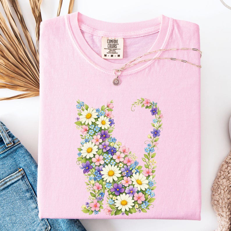 May include: A light pink Comfort Colors t-shirt featuring a floral cat design. The cat is composed of white daisies, blue, purple, and pink flowers. A delicate silver necklace with a small pendant rests on the shirt. The shirt is folded.