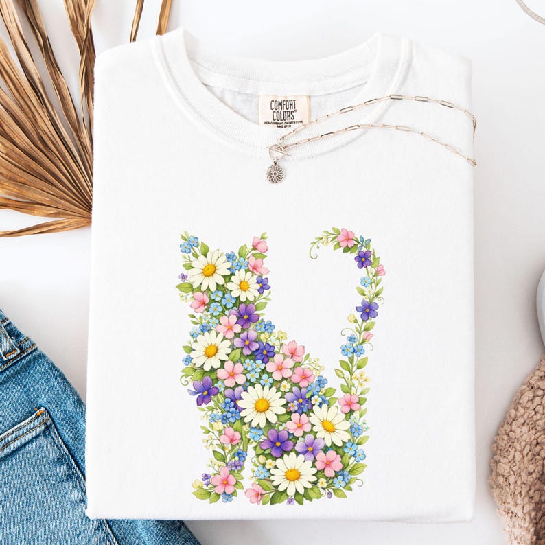 May include: White t-shirt featuring a floral cat design. The cat is composed of daisies, blue, purple, and pink flowers. The shirt has a Comfort Colors tag and a silver necklace. The shirt is folded and placed on a white surface.