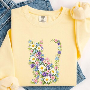 May include: A light yellow sweatshirt featuring a floral cat design. The cat is composed of white daisies, blue forget-me-nots, pink and purple flowers, and green leaves. The sweatshirt is folded on top of blue jeans.