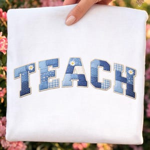 Może przedstawiać: Biała bluza z napisem „TEACH” wyhaftowanym niebieskimi literami w stylu patchwork. Każda litera ma inny niebieski wzór, w tym grochy i kratę vichy, z małymi akcentami stokrotek.