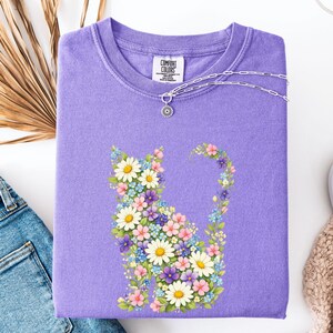 May include: A lavender Comfort Colors sweatshirt featuring a floral cat design. The cat is composed of white daisies, pink, blue, and purple flowers. A silver necklace with a flower pendant rests on the shirt. The Comfort Colors logo is visible.
