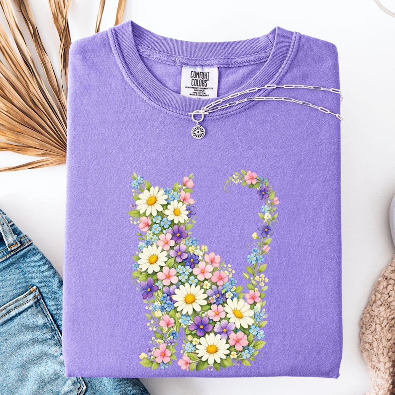 May include: A lavender Comfort Colors sweatshirt featuring a floral cat design. The cat is composed of white daisies, pink, blue, and purple flowers. A silver necklace with a flower pendant rests on the shirt. The Comfort Colors logo is visible.