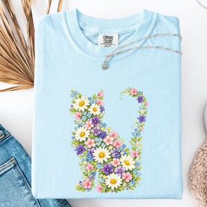 May include: Light blue Comfort Colors t-shirt featuring a floral cat design. The cat is composed of white daisies, purple, pink, and blue flowers. A silver necklace with a small pendant rests on the shirt.