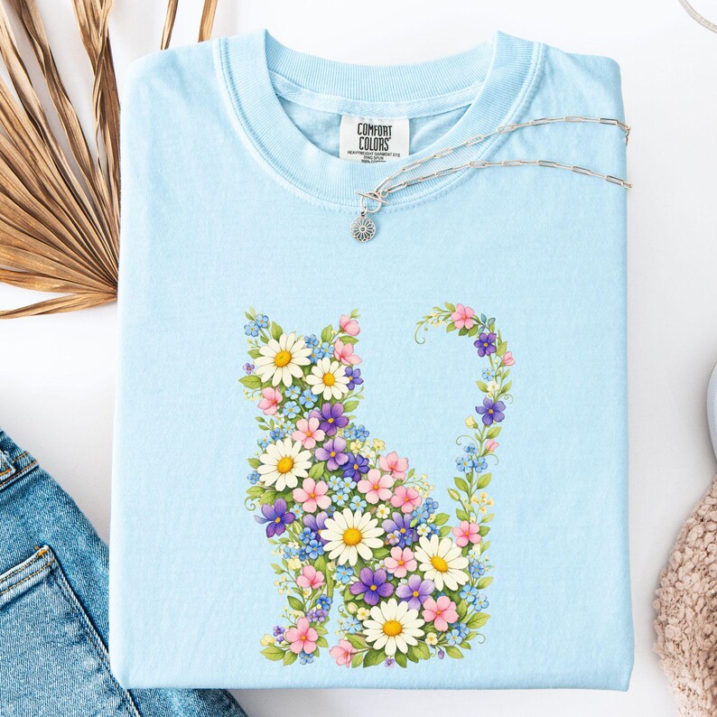 May include: Light blue Comfort Colors t-shirt featuring a floral cat design. The cat is composed of white daisies, purple, pink, and blue flowers. A silver necklace with a small pendant rests on the shirt.