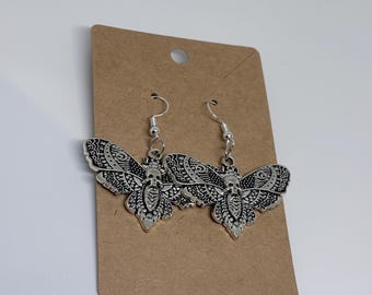 May include: A pair of silver-toned metal earrings shaped like moths with intricate filigree patterns and a skull design on the thorax. The earrings feature standard silver-coloured metal fishhook wires and are attached to a rectangular brown card.