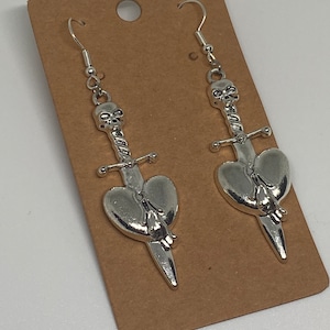 May include: A pair of silver earrings featuring a skull and dagger design with a heart pierced by the dagger. The earrings are hanging from a brown card.