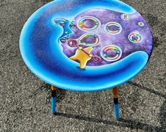 Puede incluir: Una mesa auxiliar redonda y caprichosa con un diseño de luna creciente azul que sopla burbujas. La mesa tiene un fondo morado y azul con burbujas coloridas. La mesa tiene tres patas con detalles azules, naranjas y negros.