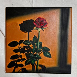 May include: A square canvas painting featuring a red rose with green leaves and stem against an orange and black background. A dark silhouette of a rose is also visible. The artwork is a still life of a flower.