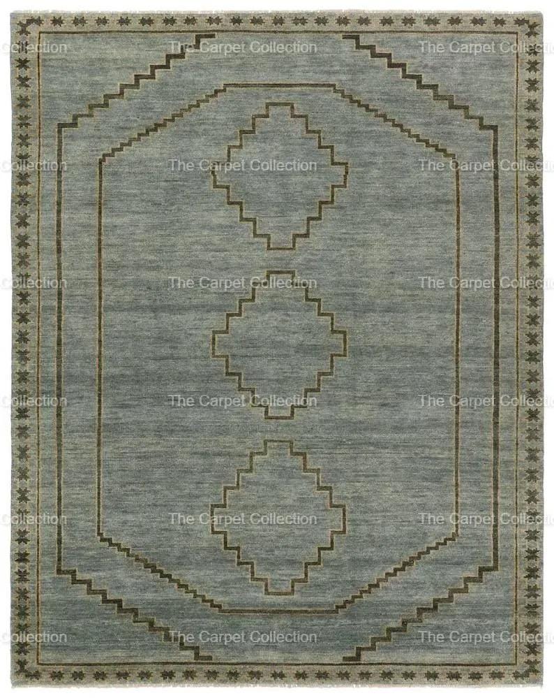 May include: A rectangular, light blue rug with a geometric design. The rug features a central pattern of diamonds and stepped lines in a darker brown color. The border is a repeating star pattern. The text "The Carpet Collection" is repeated around the edges.
