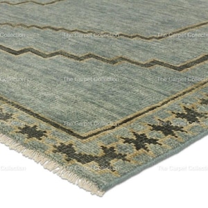 May include: Close-up of a section of a blue rug with a geometric design. The rug features a zigzag pattern in brown and a border with star-like shapes. The rug has a fringe on one edge. The text "The Carpet Collection" is visible.
