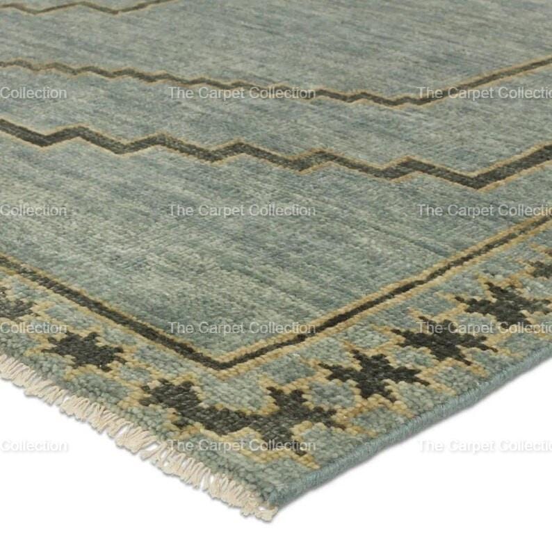 May include: Close-up of a section of a blue rug with a geometric design. The rug features a zigzag pattern in brown and a border with star-like shapes. The rug has a fringe on one edge. The text "The Carpet Collection" is visible.