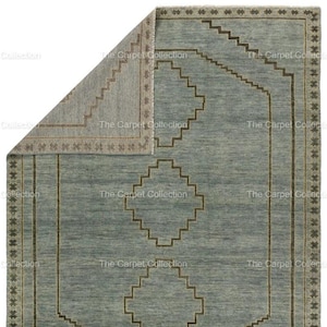 May include: A rectangular area rug with a geometric design. The rug features a light blue field with brown geometric shapes and borders. The corner of the rug is folded back, revealing a lighter, patterned underside. The text "The Carpet Collection" is visible.