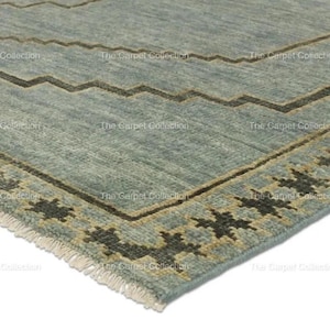 May include: A section of a blue-green rug with a geometric design. The rug features a zigzag pattern in brown and a border with star-like shapes. The rug has a fringe on one edge and the text "The Carpet Collection".