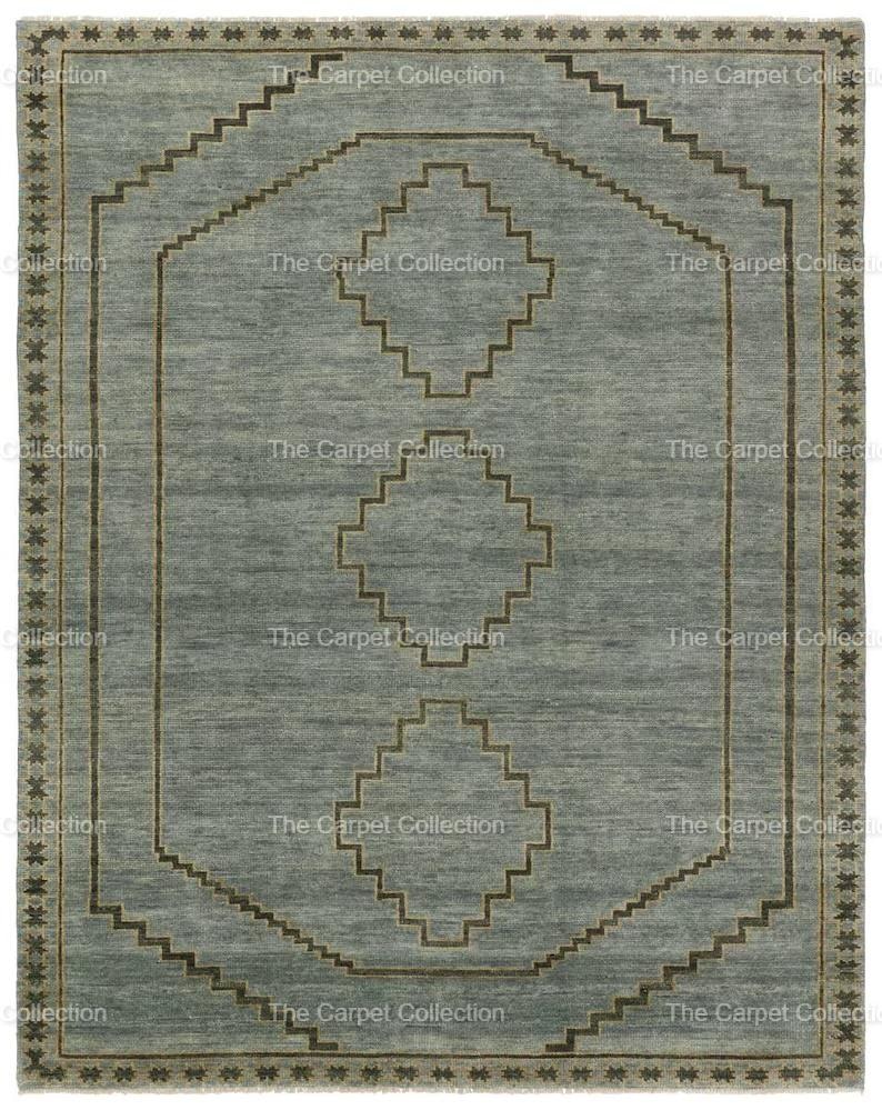 May include: A rectangular, light blue rug with a geometric design. The rug features a central pattern of diamonds and stepped lines in a darker brown color. The border has a repeating star pattern. The text "The Carpet Collection" is visible.
