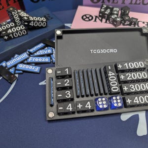 May include: A black game organizer with dice, counters, and a dice tray. The organizer has slots for dice, counters, and a dice tray. The dice are black with white numbers, and the counters are blue with white text. The text "TCG3DCRO" is visible.