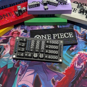 May include: A black game board with white text "ONE PIECE" and a black counter with numbers and values. Dice and other game accessories are visible, including red, green, and purple game boxes. The background features a colorful illustration.