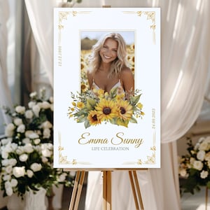 May include: A white sign with a gold frame and sunflowers. The sign reads "Emma Sunny Life Celebration" with the dates "19.11.1985" and "21.01.2023". A photo of a woman is in the center of the sign.