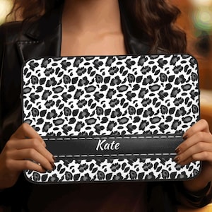 May include: A black and white leopard print laptop sleeve with a black leather band that has the name "Kate" printed on it. The sleeve is held up by a person in front of a bookshelf.