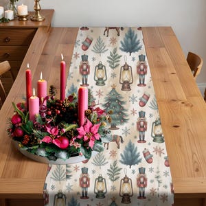 May include: A wooden dining table set for a holiday meal, featuring a festive runner with Christmas tree, lantern, and nutcracker designs. A centerpiece of pink candles and seasonal greenery adds to the holiday ambiance. Additional candles and a wooden cabinet are visible.