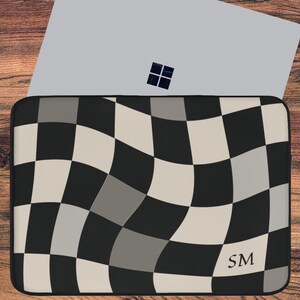 May include: Laptop sleeve with a black, white, and gray checkered pattern. The sleeve is on a wooden surface with a laptop, glasses, and a coffee cup. The initials "SM" are visible on the sleeve.