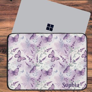 May include: A laptop sleeve featuring a repeating pattern of purple butterflies and lavender sprigs on a white background. The name "Sophia" is printed in cursive at the bottom right corner. The sleeve is black-edged and sits on a wooden surface.