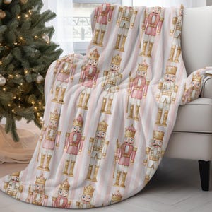 May include: A soft, pink and white striped throw blanket featuring repeating illustrations of nutcracker soldiers in pink and white uniforms with gold accents. The blanket is draped over a white armchair, near a Christmas tree.