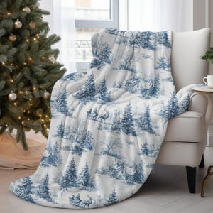 May include: A white and blue throw blanket with a winter scene design. The blanket features a repeating pattern of trees, houses, and horse-drawn sleighs. The blanket is draped over a white armchair, next to a Christmas tree.