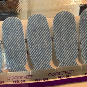 May include: Close-up of Color Street nail strips with a denim-like pattern. The nail strips are light blue with a diagonal white stripe design. The words "colorSTREET" and "PEEL OFF" are printed on the packaging.