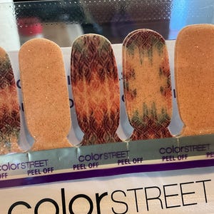 May include: Close-up of Color Street nail polish strips. The strips feature a variety of designs, including glittery peach, and patterned orange, brown, and green. The brand name "colorSTREET" is visible at the bottom of the image.
