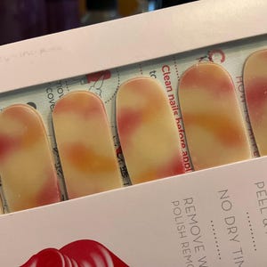 May include: A package of nail polish strips. The strips have a cream base with blended orange and red hues. The package is pink and has text that reads "PEEL & APPLY" and "NO DRY TIME".