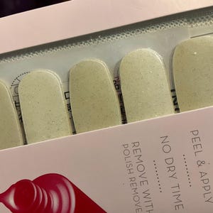 May include: A package of light green nail polish strips with a glittery finish. The package is open, revealing several strips. Text on the package reads "PEEL & APPLY" and "NO DRY TIME".