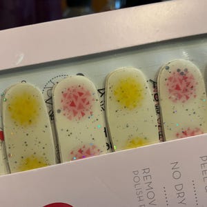 May include: A set of nail polish strips with a glittery, speckled finish. Each strip features a pastel yellow, pink, or blue geometric design. The strips are packaged in a white box with text.
