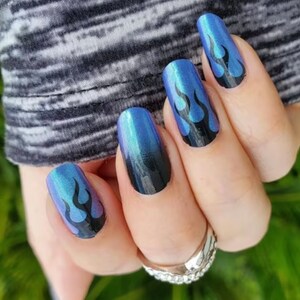 May include: Close-up of a hand with blue and black ombre nail polish with black flame designs on each nail.